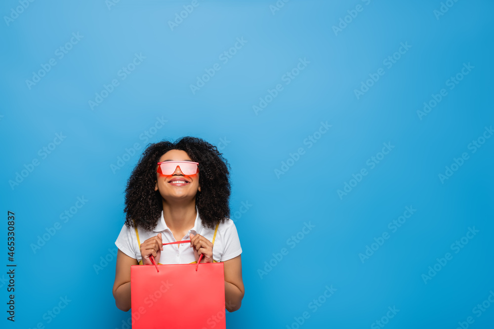 cheerful african american woman in fashionable eyeglasses holding shopping bag isolated on blue cheerful african american woman in fashionable eyeglasses holding shopping bag isolated on blue