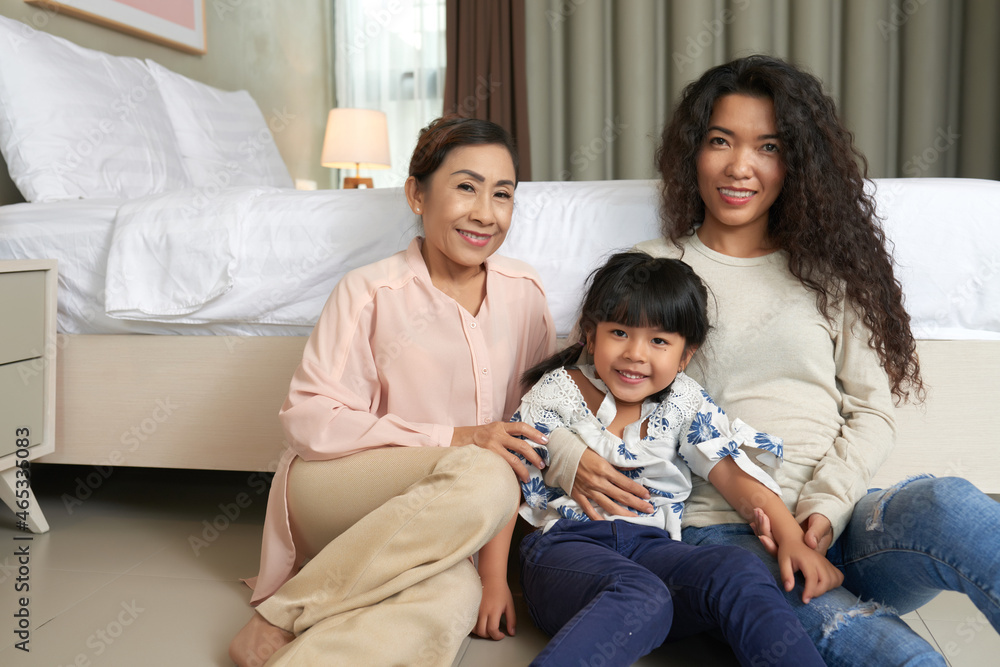 Portrait of smiling cute Asian girl sitting in hugs of mother and grandmother against bed Portrait of smiling cute Asian girl sitting in hugs of mother and grandmother against bed