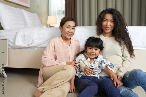 Portrait of smiling cute Asian girl sitting in hugs of mother and grandmother against bed