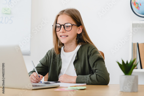 Smart caucasian schoolgirl student pupil learning online, using laptop, listening to lessons from home, doing homework on lockdown