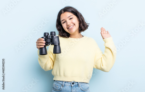 pretty young woman smiling happily, waving hand, welcoming and greeting you. binoculars concept photo