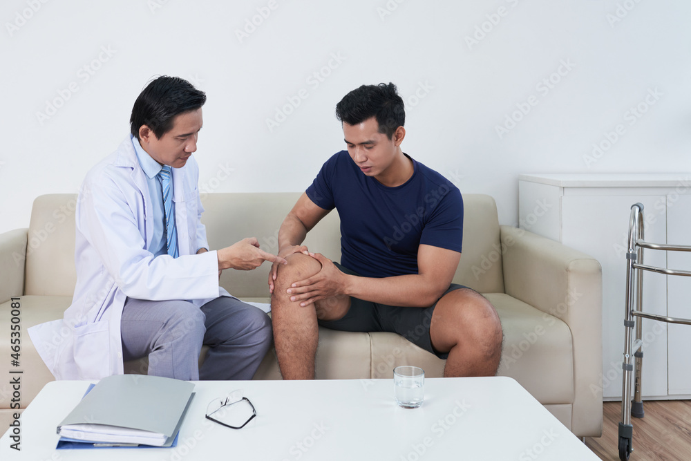 Horizontal medium long shot of young Asian man sitting on sofa in doctors office having consultation about pain in his knee Horizontal medium long shot of young Asian man sitting on sofa in doctors office having consultation about pain in his knee