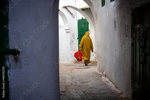 In the medina of Chefchaouen in Morocco. A man wearing the traditional Berber coat walks down the narrow street carrying a red bag photo