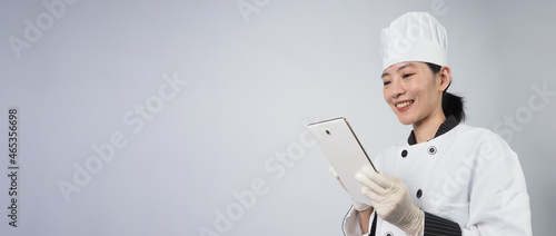 Asian woman chef holding smartphone or digital tablet and received order from online shop or merchant application. she smiling in chef uniform and standing in white background. Online food merchant. 