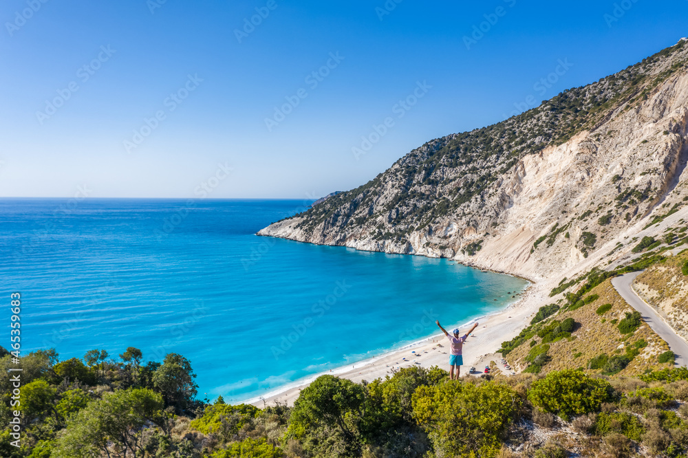 Man tourist standing on top of a rock, raising hands with an exciting feeling of freedom, looking at Myrtos Beach. Cephalonia island, Greece
