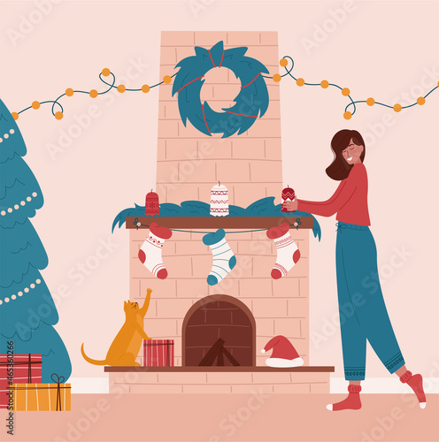 Cute girl decorating fireplace in living room.Woman getting ready for Christmas eve. Fireplace with Christmas decorations: socks, candles, gifts, wreath, garland.Funny cat trying to catch socks. 