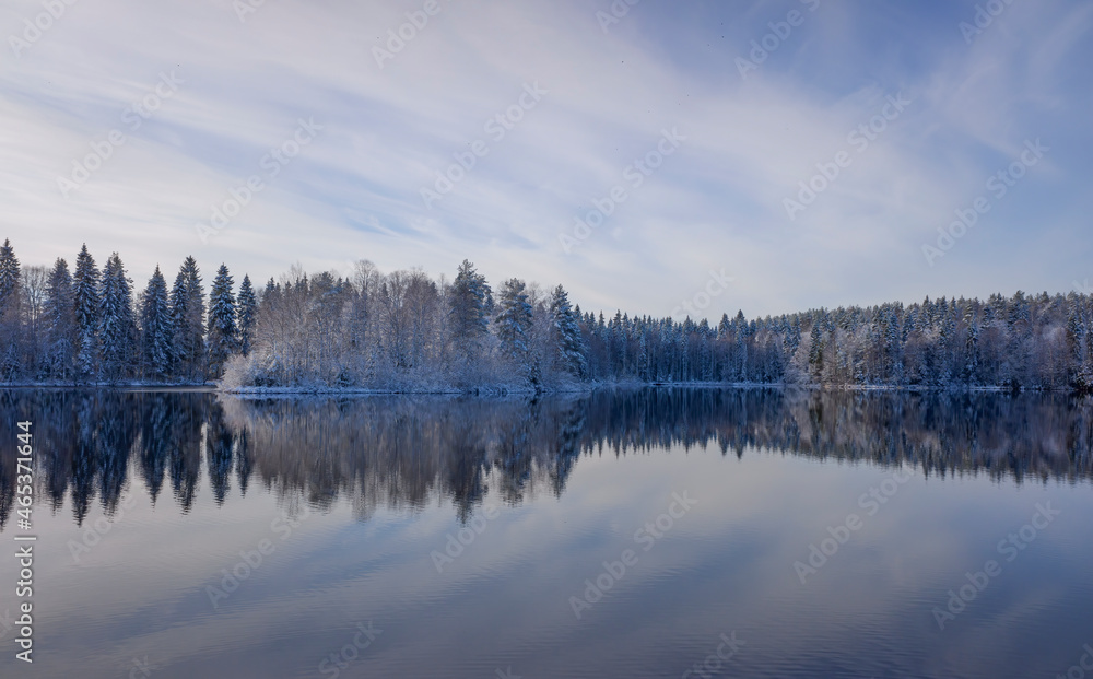 a snow-covered forest with the first snow, a reflection in the lake against the background of a blue sky with clouds