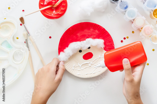 The child glues the parts Santa made from paper plate. Handmade. Project of children's creativity, handicrafts, crafts for kids. Preparation for christmas. Decoration.