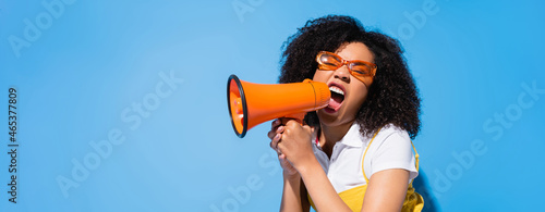 excited african american woman in eyeglasses shouting in loudspeaker on blue, banner photo