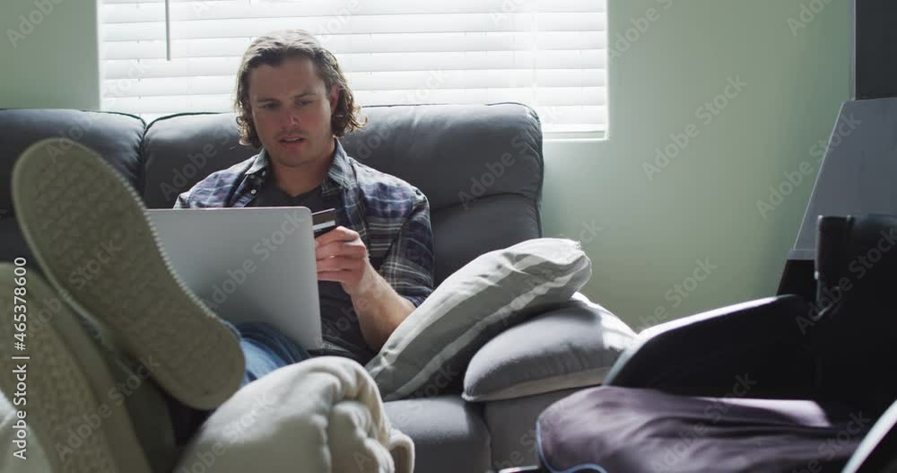 Happy caucasian disabled man sitting on sofa in living room holding card and using laptop Happy caucasian disabled man sitting on sofa in living room holding card and using laptop