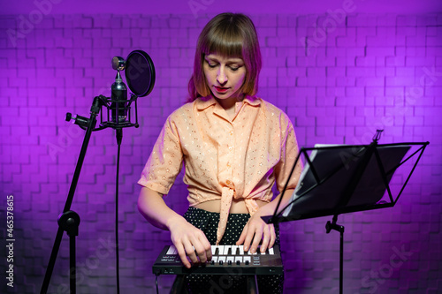 young caucasian singer watches and controls the keys in front of the music stand, microphone with a pop filter on a tripod in a studio with a soundproof wall and purple backlight