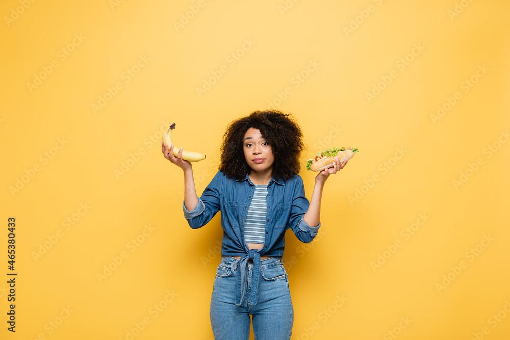 confused african american woman looking at camera while choosing between bananas and hot dog on yellow confused african american woman looking at camera while choosing between bananas and hot dog on yellow