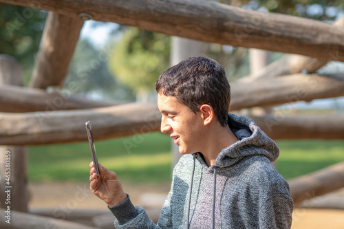 young brown-haired boy looking at his cell phone in a park wearing a gray hooded sweatshirt