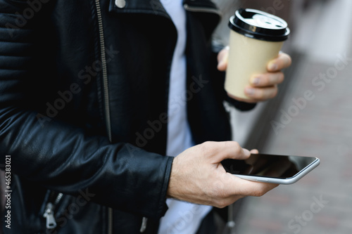 Cropped view of man in leather jacket holding smartphone with blank screen and takeaway coffee outdoors 