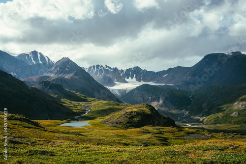 Atmospheric alpine landscape with mountain lake in green valley and glacier under cloudy sky. Awesome highland scenery with beautiful glacial lake among sunlit hills and rocks against mountain range.