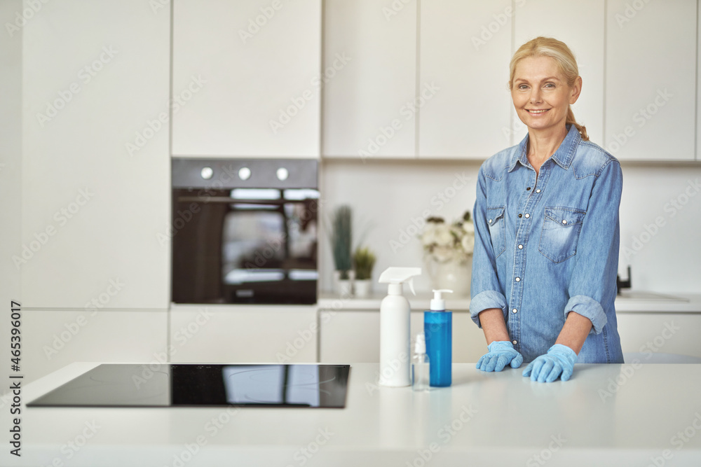 Smiling woman in rubber gloves ready for clean up in the kitchen. Detergents, sanitizer spray and gel in bottle on the surface Smiling woman in rubber gloves ready for clean up in the kitchen. Detergents, sanitizer spray and gel in bottle on the surface