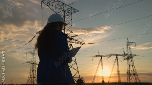 Engineer woman power engineer in white helmet checks power line using computer tablet. Woman Electricity supply company employee works outdoors, services high voltage electric lines at sunset