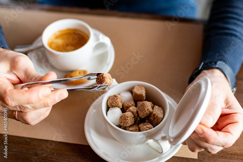 Hand puts a sugar cube in the coffee cup.A man's hand uses a cutlery to take a lump of sugar to add to black coffee.