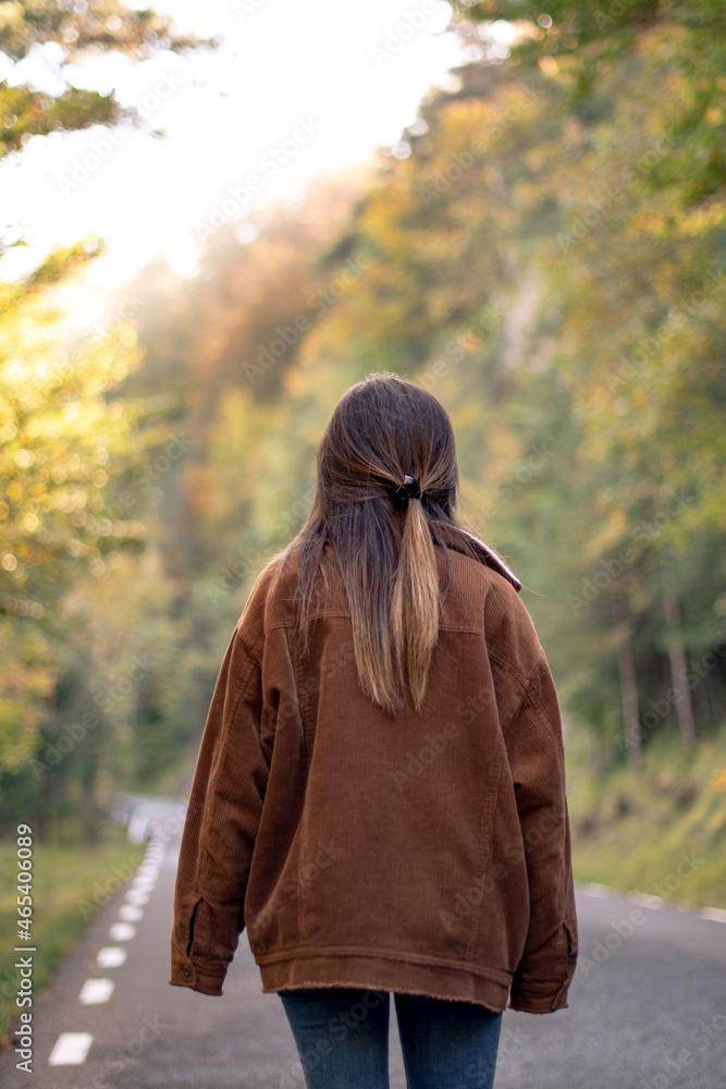 back girl walking with a brown coat in autumn in a road to a green and brown trees forest