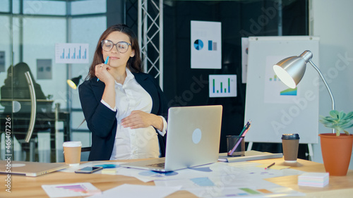 Beautiful Young Pregnant Woman at Work in a Comfortable Modern Office. A Happy, Attractive, Pensive Office Worker at the Workplace. Motherhood, Work, and Comfort.