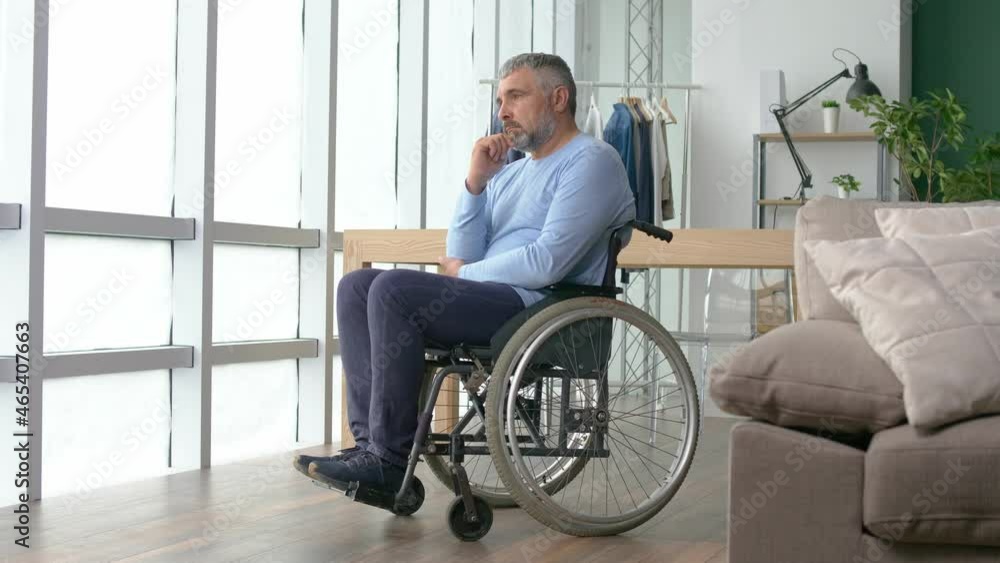 Depressed Lonely Middle-aged Man Looking Out the Window, a Frustrated Mature Man Sitting in a Wheelchair at Home Alone. Privacy, Care, Retirement, Disability. Depressed Lonely Middle-aged Man Looking Out the Window, a Frustrated Mature Man Sitting in a Wheelchair at Home Alone. Privacy, Care, Retirement, Disability.