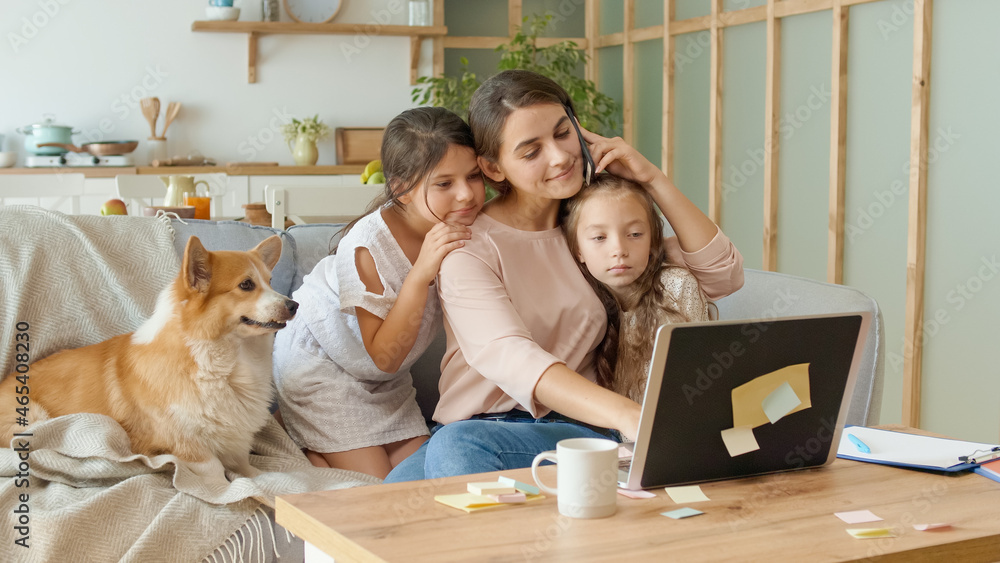 Lovely Mother Working Remotely From Home While Hugging Kids are Sitting Beside on the Couch. Mother is Stroking and Kissing Her Little Daughters During Working. A Busy Happy Mother Working Remotely