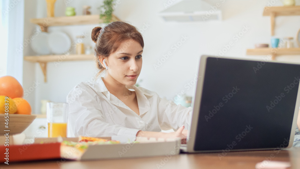 Pretty Woman Sitting at the Kitchen and Work Remotely. Woman Put Her Legs on the Table, on the Table Lying Pizza, Fresh Juice, and Notebooks.Attractive Woman Sitting With Headphones at the Bright Room