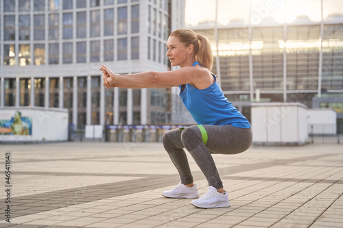 Attractive middle aged woman in sportswear doing squats using resistance band, having workout in the city