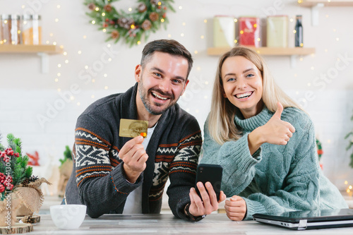 Young happy family during the New Year and Christmas holidays at home joyful buy gifts online using a smartphone and credit card