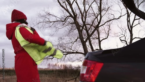 The woman takes a yellow vest out of the trunk of the car and puts it on.