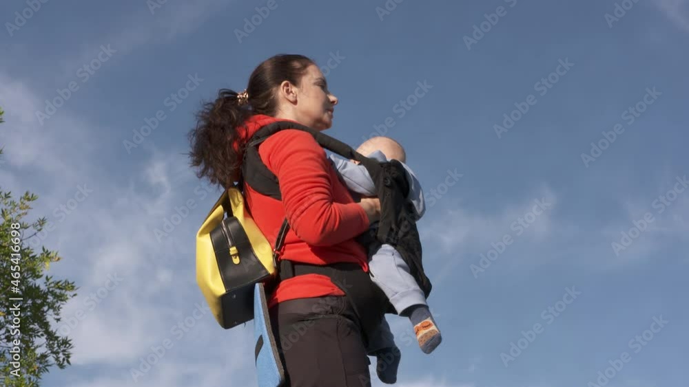 Mother carrying her little baby in infant kid carrier against blue sky, family vacations adventure, mom with baby boy hiking, summer active lifestyle. High quality 4k footage Mother carrying her little baby in infant kid carrier against blue sky, family vacations adventure, mom with baby boy hiking, summer active lifestyle. High quality 4k footage