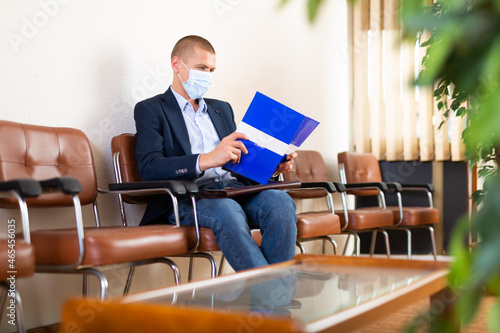 Stylish successful man in face mask with briefcase waiting for job interview in office lobby