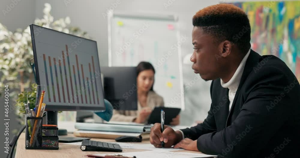 A dark-skinned man in a suit sits at a desk in front of a computer monitor, describes graphs printed on paper, compares them with the diagrams on the screen, signs them, confirms their conformity A dark-skinned man in a suit sits at a desk in front of a computer monitor, describes graphs printed on paper, compares them with the diagrams on the screen, signs them, confirms their conformity