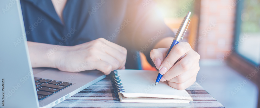 Business woman hand writing on a notepad with a pen and use a laptop computer in the office.