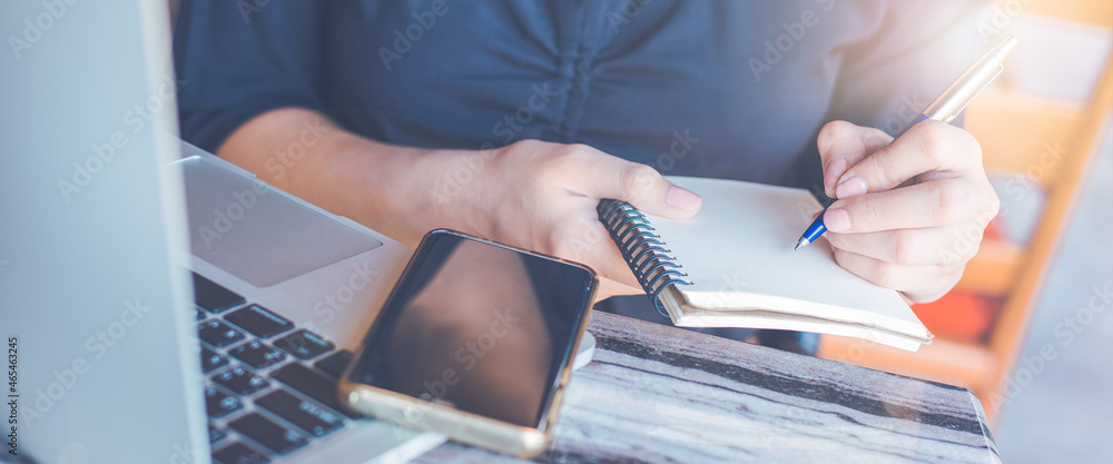 Business woman hand writing on a notepad with a pen and use a laptop computer in the office.