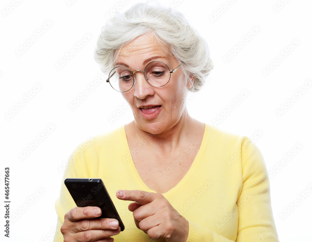 Pretty senior female in casual outfit using modern smartphone and laughing out loud while standing on white background. Old woman watching funny videos on internet. Pretty senior female in casual outfit using modern smartphone and laughing out loud while standing on white background. Old woman watching funny videos on internet.