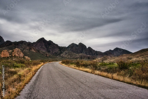 Fényképezés View of a road asphalt way between wild grass and black high rocky mountains und