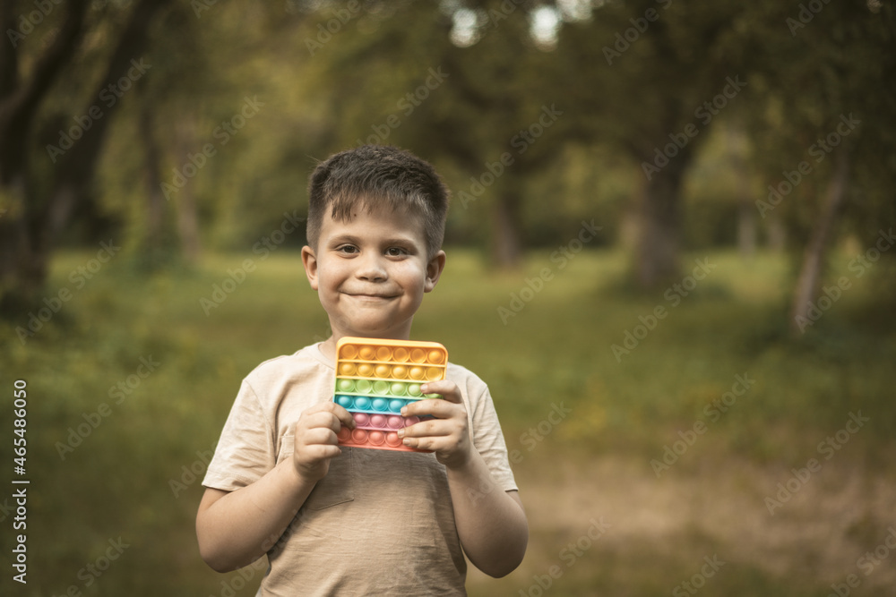 Smiling Boy With Rainbow Pop It Walks in the Park. Nice Kid Plays Pop It Outdoor. High quality photo Smiling Boy With Rainbow Pop It Walks in the Park. Nice Kid Plays Pop It Outdoor. High quality photo