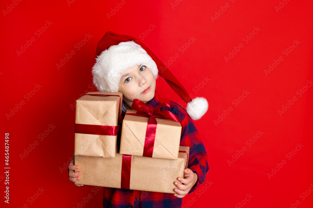 Happy holiday and Merry Christmas. Portrait of a boy in a cap with gifts in his hands on a red background. A place for text. Happy holiday and Merry Christmas. Portrait of a boy in a cap with gifts in his hands on a red background. A place for text.