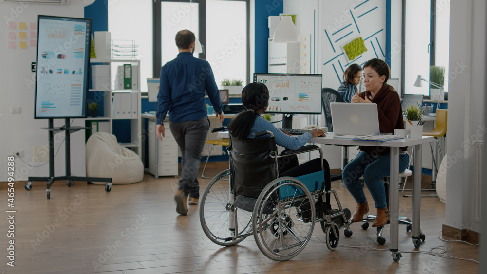 Multiethnic team working at financial project in startup financial company workplace, black immobilized woman in wheelchair and coworker discussing solution for project.