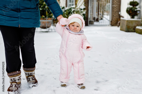 Adorable little baby girl making first steps outdoors in winter through snow. Cute toddler learning walking. Mother holding child on hand. Daughter and mum walk together. Happy family