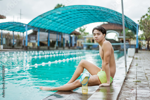 Boy teen swimmer in swimming trunks sitting resting with drinking bottle