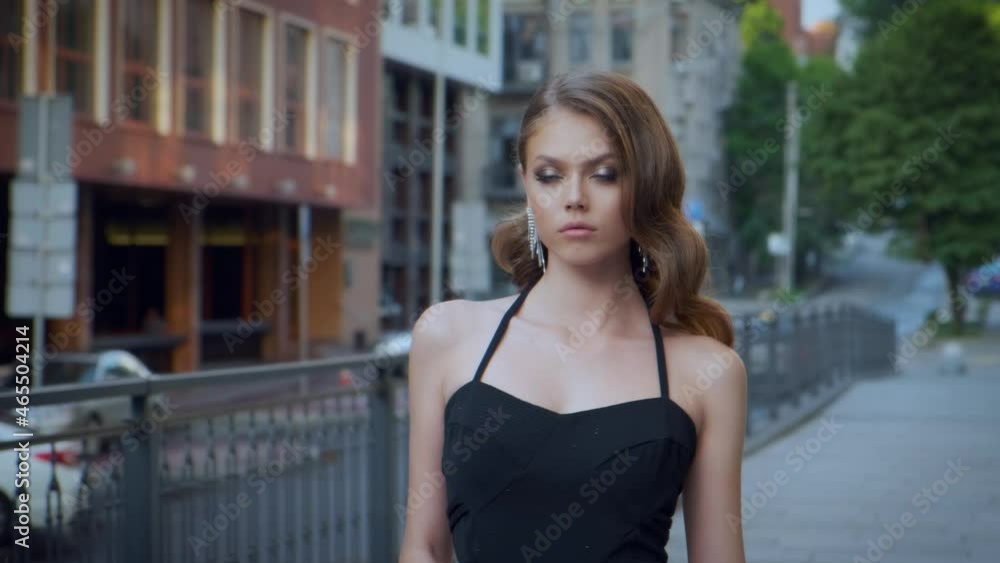 Beautiful sexy fashion woman walking along street summer modern city. Brunette girl, long wavy hair. Black luxury stylish classic elegant dress. Evening make-up. Sparkle earrings. Green trees sidewalk Beautiful sexy fashion woman walking along street summer modern city. Brunette girl, long wavy hair. Black luxury stylish classic elegant dress. Evening make-up. Sparkle earrings. Green trees sidewalk