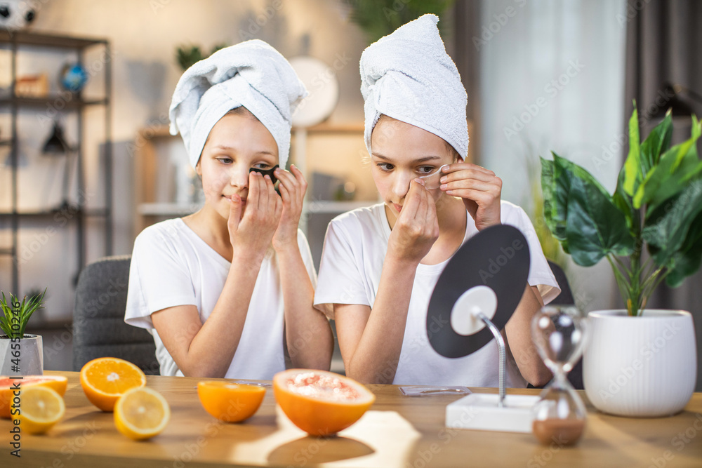 Two caucasian sisters wearing towel on head and white t-shirt sitting together at desk and applying patches under eyes. Beauty procedures for keeping youth.