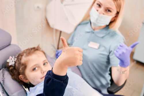Focus on girl s hand giving thumbs up while woman dentist in medical mask sitting beside kid  showing approval gesture after dental procedure. Concept of pediatric dentistry and dental care approval.