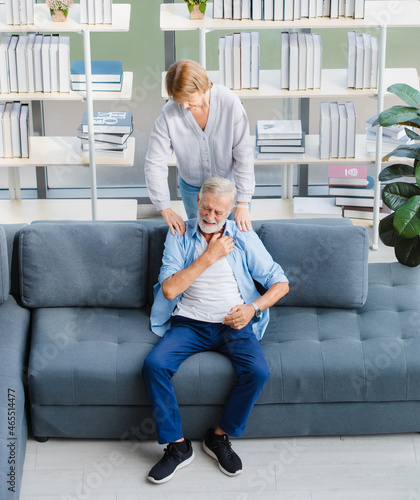 Lovely caucasian senior elderly wife standing massage happy white hair and bearded husband shoulder sitting relaxing resting on cozy sofa in living room at home in front of bookshelf and tree pot