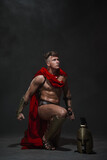 A young, athletic man dressed as a Roman warrior in a red cloak and helmet on a black background dropped to one knee.