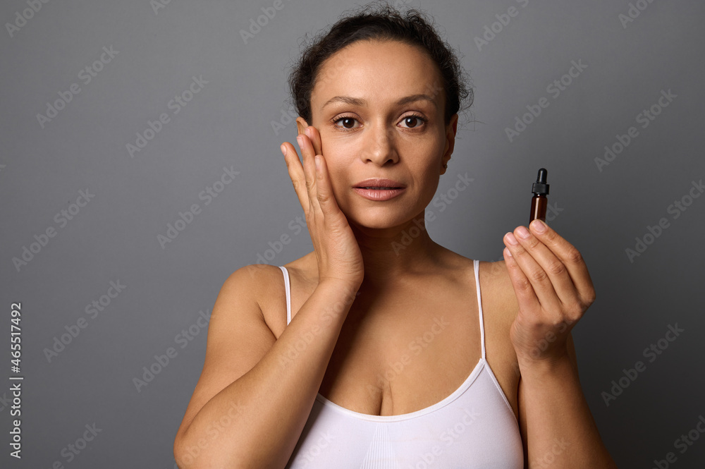 Beautiful middle aged woman holds a jar with beauty product, applies it on her face and performs anti-aging lymphatic drainage massage for rejuvenation and skin smoothing. SPA, cosmetology concept Beautiful middle aged woman holds a jar with beauty product, applies it on her face and performs anti-aging lymphatic drainage massage for rejuvenation and skin smoothing. SPA, cosmetology concept