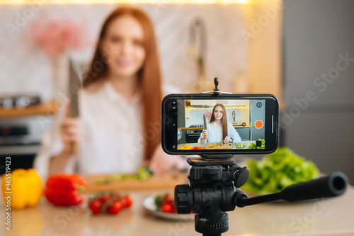 Display of camera recording video blog for food blogger woman cutting vegetable in modern kitchen talking about healthy vegan eating. Influencer vlogger girl live streaming nutrition masterclass.