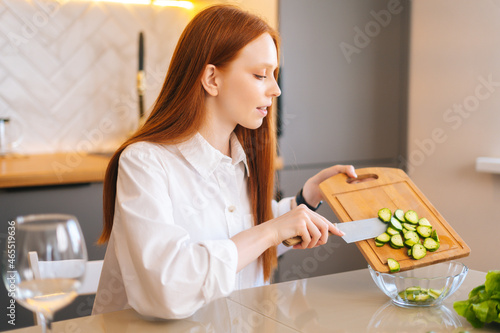 Side view of attractive young redhead woman throwing sliced cucumbers in glass bowl sitting at table in kitchen room. Happy female cooking vegetarian dieting salad full of vitamins.